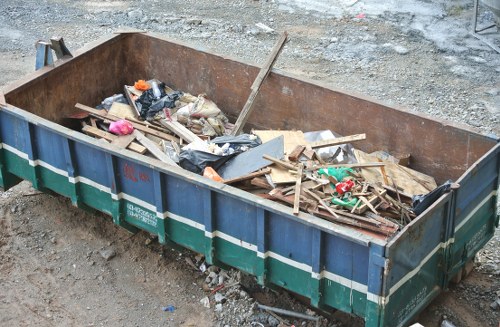 A large, rectangular metal skip with a rusted, brownish surface and a green and blue striped exterior, positioned on a gravel surface outdoors. Inside the skip, there are various types of waste materials including wooden planks, cardboard, black plastic bags, and miscellaneous debris, some of which are partially crushed or bent. The wooden planks are light brown with rough textures, while the plastic bags are glossy and black. The gravel surrounding the skip appears uneven, with small stones and dirt visible, indicating a site area potentially in Merton. The background shows a continuation of the gravel area with no other objects or structures clearly visible, emphasizing the domestic or construction waste typical of rubbish clearance services provided by Merton House Clearance.