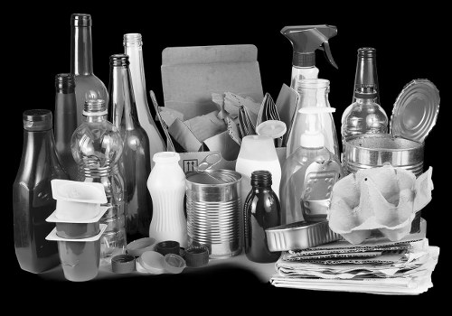 A collection of various empty glass bottles, plastic containers, and packaging materials arranged on a surface, with some bottles standing upright and others leaning or stacked. Among the items are clear glass bottles with narrow necks, plastic spray bottles, and some metallic cans and foil wrappers. To the right, there are crumpled brown paper or cardboard packaging and a partially visible cardboard box, suggesting a collection of household waste or recyclables. The background appears dark, highlighting the reflective surfaces of the bottles and containers, and the scene is typical of waste items prepared for rubbish removal, reflective of services provided by Merton House Clearance in the Merton area.