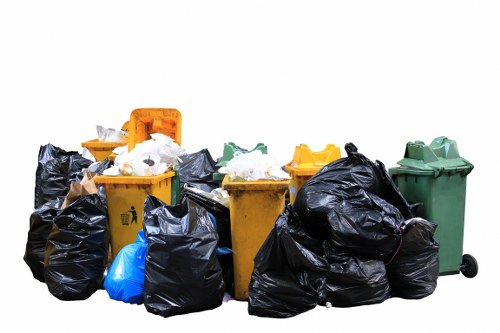A collection of multiple rubbish bins and large black and blue garbage bags are positioned outdoors on a paved surface, with a white background. The bins are of various colours including yellow, green, and orange, made of plastic with textured surfaces, and have lids, some of which are open or partially lifted. The black waste bags appear full and are loosely stacked, with some leaning against the bins, and others placed directly on the ground. The yellow bins show signs of wear, and the green bin has small wheels at its base. The scene suggests a waste collection or clearance operation typical of domestic or light commercial rubbish removal, possibly in a residential area within the vicinity of Merton, as associated with Merton House Clearance. The overall setting emphasizes the practical and neutral nature of rubbish disposal tasks, with no visible clutter or other environmental features, focusing solely on the waste containers and bags prepared for removal or disposal.
