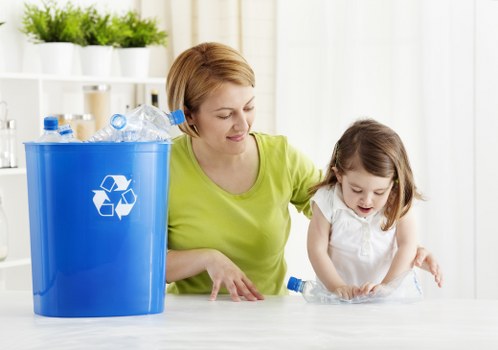 A woman with short, light brown hair, wearing a lime-green top, is sitting at a white table with a young girl who has medium-length brown hair and is dressed in a white top. They are engaged in a recycling activity, with the woman helping the girl sort or place plastic bottles into a bright blue recycling bin positioned on the left side of the image. The bin features a white recycling symbol and is partially filled with clear plastic bottles and caps. The background shows a white kitchen setting with open shelving, white storage jars, and potted plants, creating a clean, organized atmosphere. The scene captures a moment of eco-friendly responsibility, emphasizing waste separation or rubbish management within a domestic environment, and subtly linking to rubbish removal services in Merton or surrounding areas.